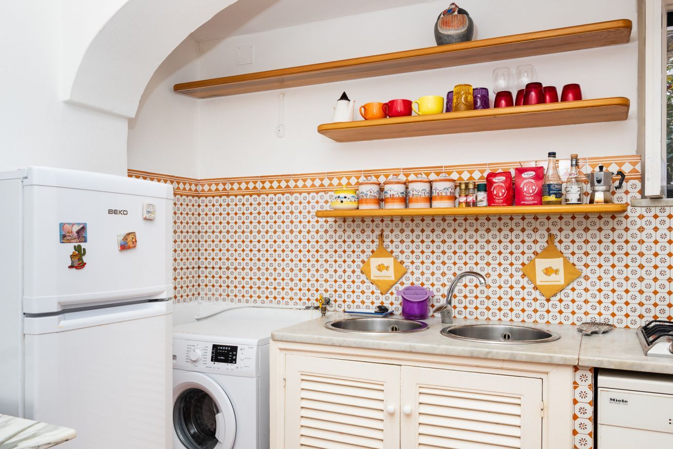 A kitchen with a washing machine, a refrigerator, wooden shelves with cups, and patterned backsplash.