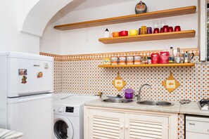 Eine Küche mit Waschmaschine, Kühlschrank, Holzregalen mit Tassen und einer strukturierten Spritzschutzwand.