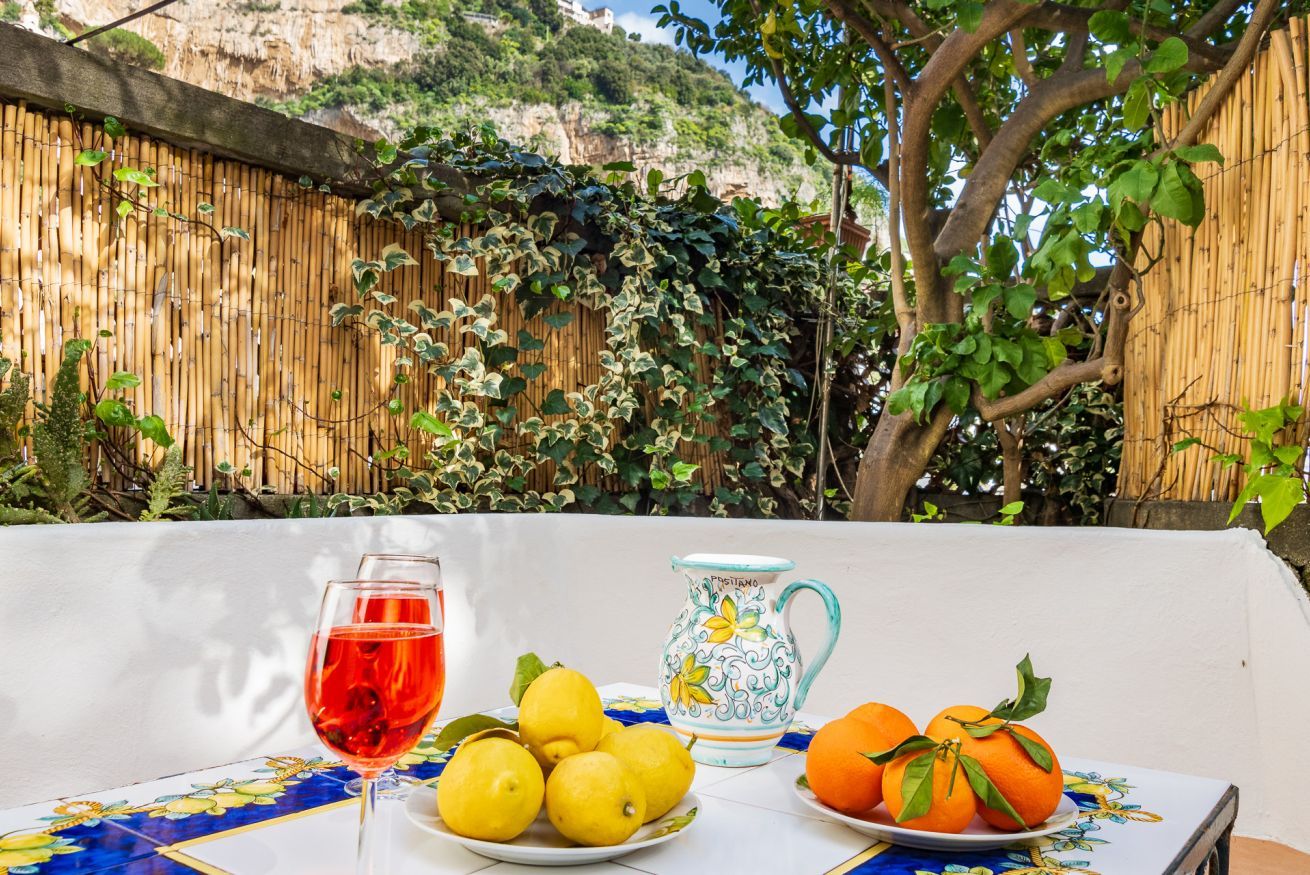 Una mesa con un vaso de bebida roja, limones, naranjas, una jarrón y un plato decorativo, frente a una valla de bambú y un árbol.