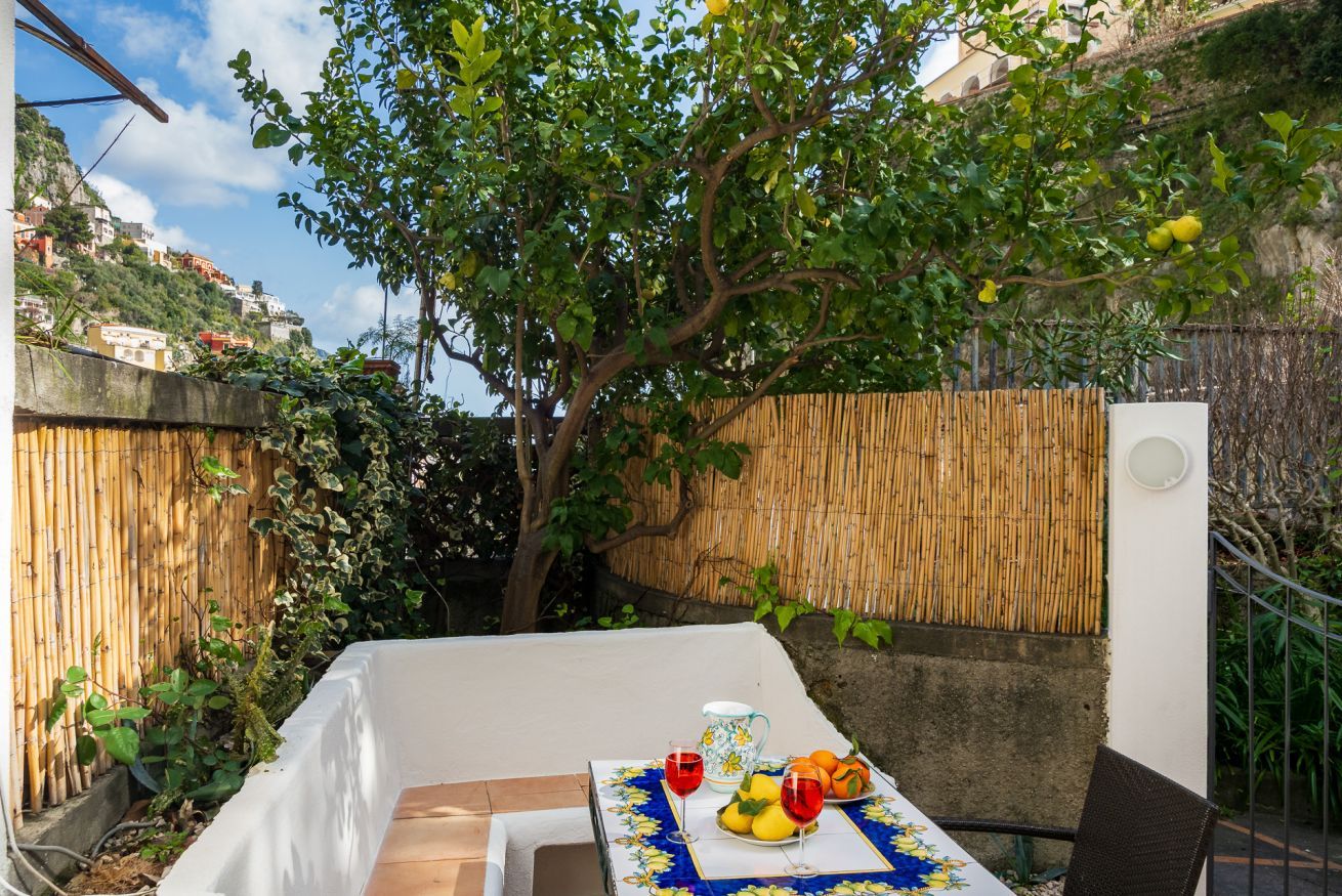Una mesa con mantel, una jarrón, vasos y fruta en un jardín.