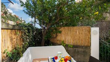 Una mesa con mantel, una jarrón, vasos y fruta en un jardín.
