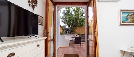 Quarto com televisão sobre um móvel branco, porta que dá para área externa com mesa e cadeiras, e piso estampado.