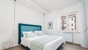 Ein Schlafzimmer mit einem türkisblauen Kopfteil, weißer Bettwäsche, einer geometrischen Stehlampe und einem Fenster mit weißen Vorhängen.
