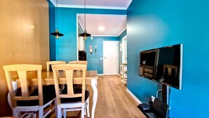 Un coin repas avec des chaises et une table en bois, un téléviseur fixé au mur et une porte visible au fond.