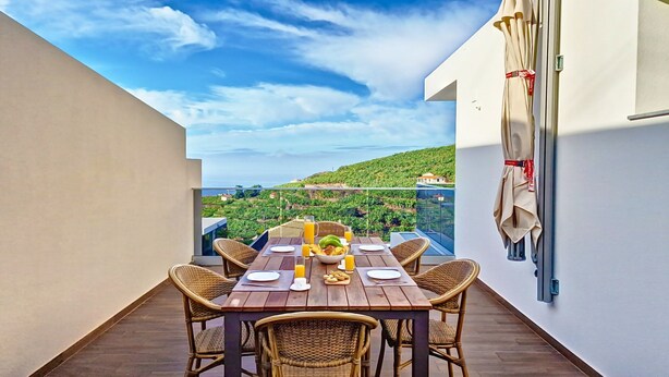 A balcony with a wooden table set for two, wicker chairs, and a view of a green landscape. - Allure Alannas Villa by LovelyStay (Funchal)