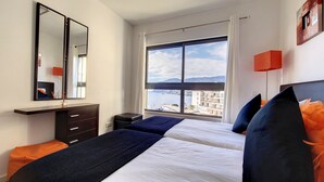 A hotel room with a large window, a bed with blue and white bedding, a dark wooden dresser, and a mirror. - Sunset Bay by LovelyStay (Funchal)