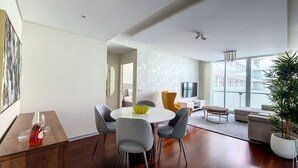 Un salon moderne avec une table à manger ronde, des chaises grises, un canapé et une grande fenêtre donnant sur des immeubles.