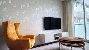Un salon moderne doté d’une grande fenêtre, d’un téléviseur à écran plat fixé au mur, d’un fauteuil jaune, d’une console blanche et d’une table basse ronde en bois.