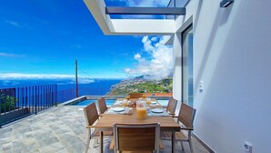A dining area with a table set for four, overlooking a coastal landscape with a mountain in the distance. - Ohana Ocean View by LovelyStay (Arco Da Calheta)