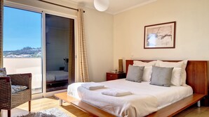 A bedroom with a large bed, a nightstand, a wicker chair, and a view through a sliding door. - Lucy Sweet Home by LovelyStay (Canico)