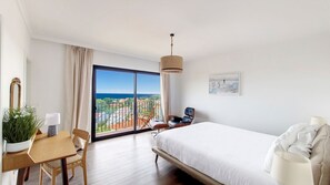 Ein modernes Hotelzimmer mit einem großen Bett, einem Schreibtisch mit Stuhl, einer Zimmerpflanze und Blick auf Stadt und Meer durch raumhohe Fenster.