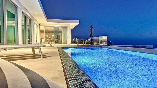 Un espace piscine moderne doté d’une piscine à l’eau d’un bleu limpide, d’un coin salon abrité et d’une salle à manger fermée par des baies vitrées.