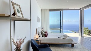 Une chambre moderne avec un grand lit, une table de chevet, une chaise et une vue sur l’océan grâce à de grandes fenêtres du sol au plafond.