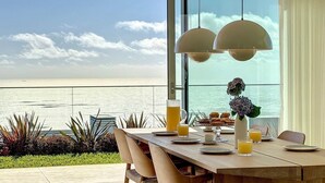 Une table à manger est dressée avec des assiettes, des verres et une carafe de jus d’orange. De grandes fenêtres offrent une vue sur une plage, avec l’océan et le ciel en toile de fond.