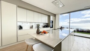Une cuisine moderne dotée d’un grand îlot central, de meubles blancs et offrant une vue sur l’océan.