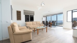 Un salon moderne avec un canapé, un fauteuil et une table basse en verre. On y trouve également un coin repas avec des chaises et une table, ainsi qu’une grande fenêtre offrant une belle vue.