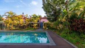 A swimming pool with a wooden deck, surrounded by tropical plants and a building with a red roof.
