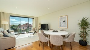 A modern living room with a large sliding glass door, a round dining table with six chairs, a flat-screen TV, and a large potted plant.