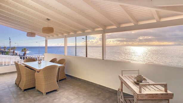 Een ruim balkon met uitzicht op de oceaan, rieten meubels en een houten kar.