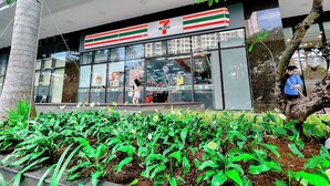 A 7-Eleven store with a modern glass facade and a well-maintained garden area in front. - Helios Rivergate Apartment (Ho Chi Minh City)