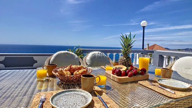 Ein Frühstücksbuffet mit Croissants, Früchten und Saft auf einem Balkon mit Blick auf das Meer.