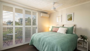 Une chambre à coucher comprenant un lit, un ventilateur de plafond, un appareil de climatisation, une fenêtre avec des stores et une petite table de chevet avec une lampe et des objets décoratifs.