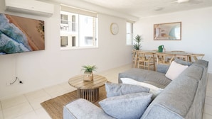 Un salon avec un canapé gris, une table basse en bois ornée d’une plante, une table à manger avec des chaises, un téléviseur fixé au mur et un ventilateur de plafond.