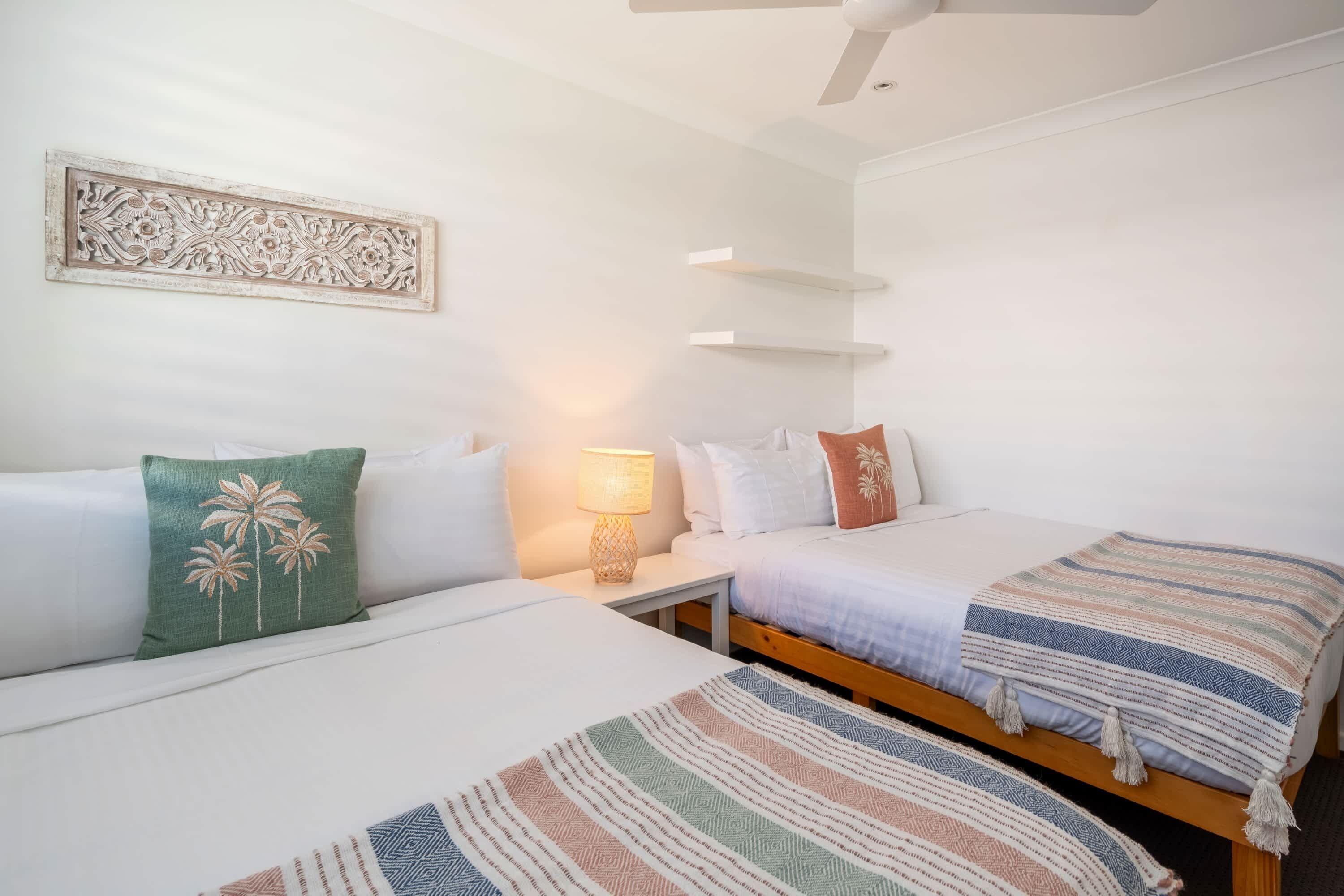 Quarto de hotel com duas camas, uma mesa de cabeceira com abajur, um ventilador de teto e um objeto decorativo na parede.