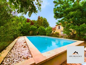 A swimming pool with a pebble border, surrounded by lush greenery and a house in the background. - Gîte de la Draillette (Noves)