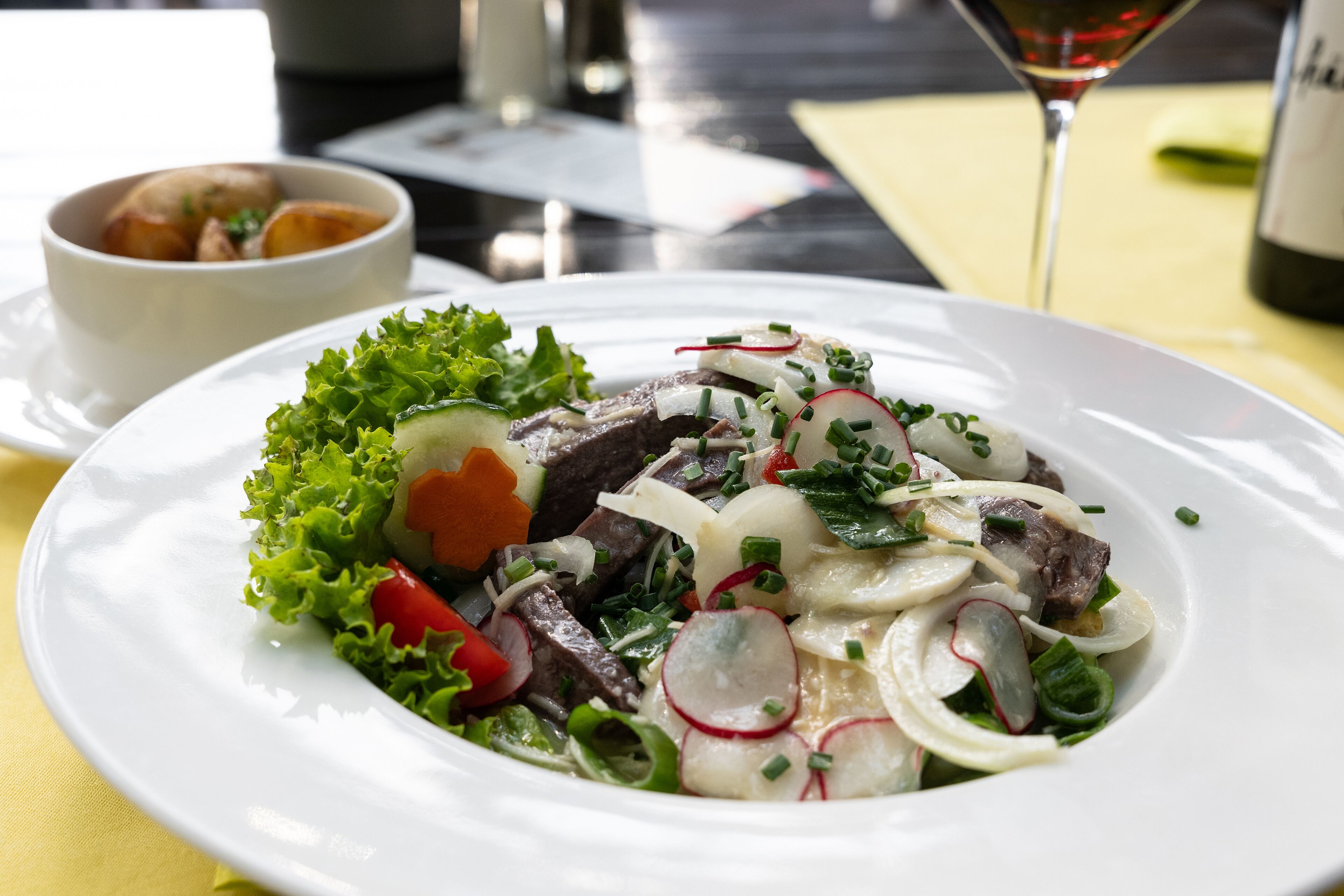 A white plate with a salad containing lettuce, radishes, and meat, accompanied by a small bowl of bread.