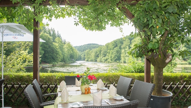 Un espace de restauration en plein air, avec des chaises en osier et une table dressée pour deux, offrant une vue sur une rivière et des collines boisées.