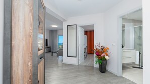 A hotel room with a wooden floor, a large window, a flat-screen TV, a dining area, and a bathroom with a glass shower.