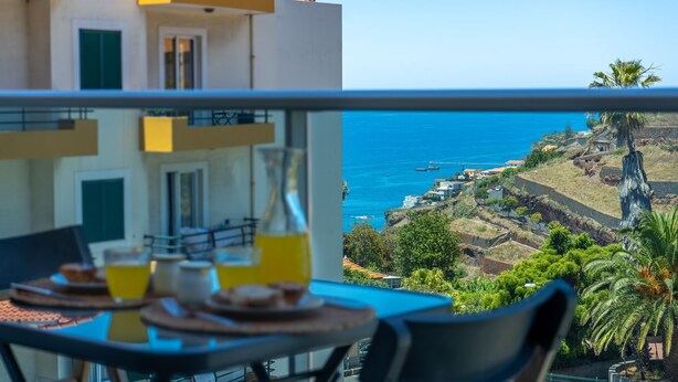 A balcony with a table set for breakfast, overlooking a coastal landscape with buildings and a clear sky.