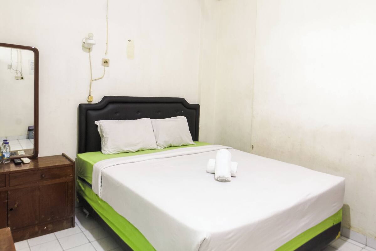 Habitación de hotel con una cama individual, una mesita de noche de madera, un espejo y una colcha verde y blanca.