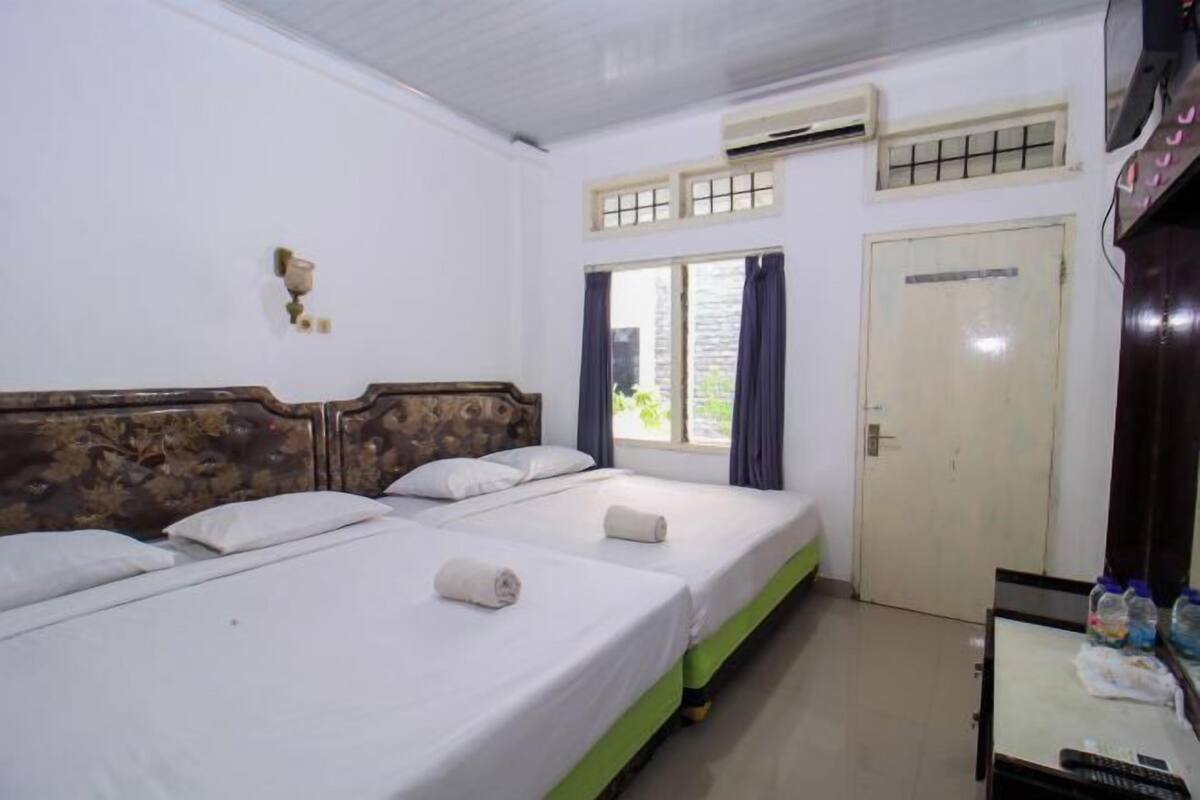 Habitación de hotel con dos camas, cabecera de madera, ventana con cortinas, equipo de aire acondicionado y una puerta.