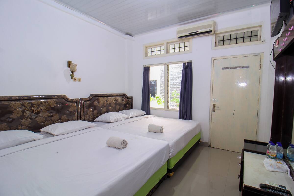 Habitación de hotel con dos camas, cabecero de madera, ventana con cortinas, equipo de aire acondicionado y una mesita con un control remoto.