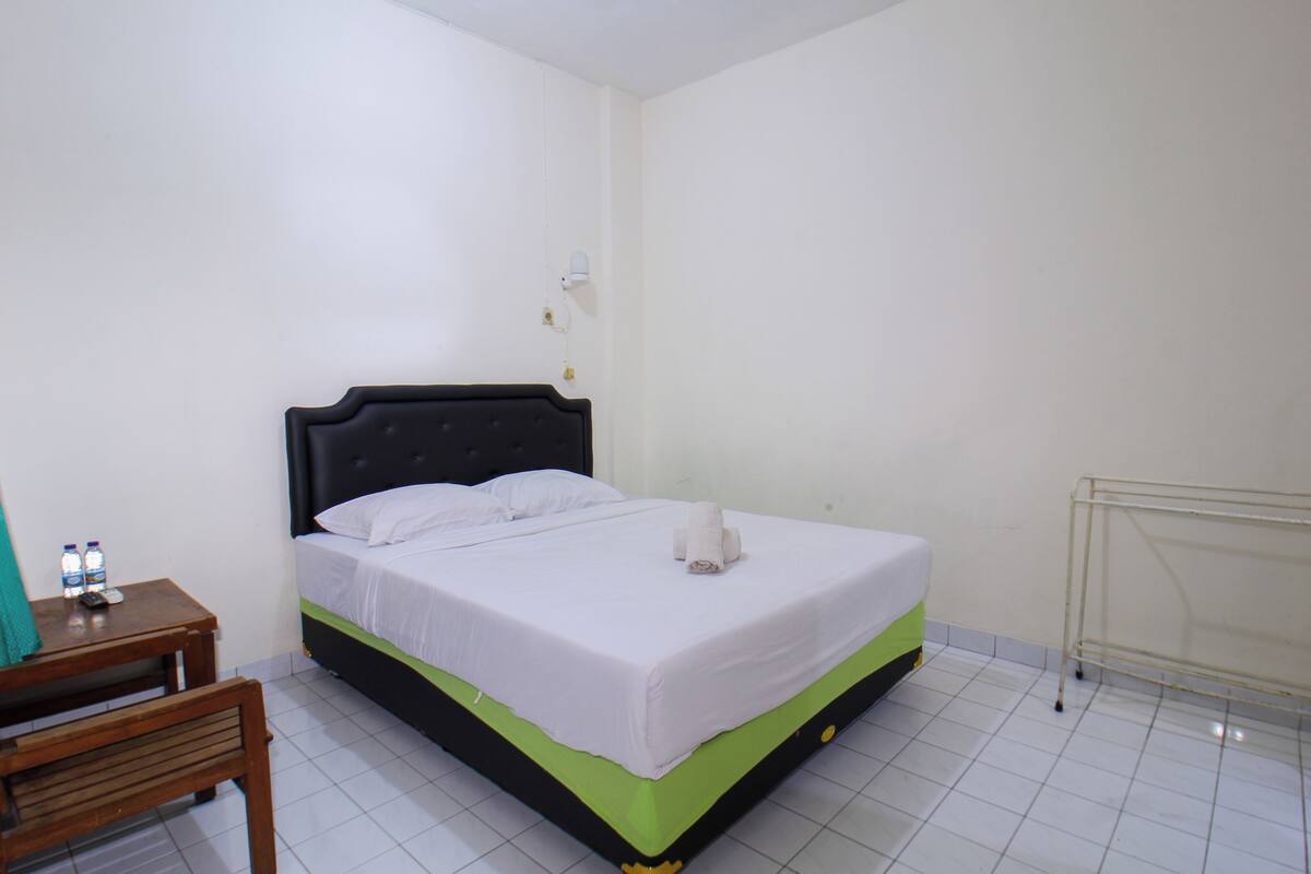 Una cama individual con estructura verde y negra, ropa de cama blanca y una mesita de noche pequeña con un control remoto y agua embotellada.