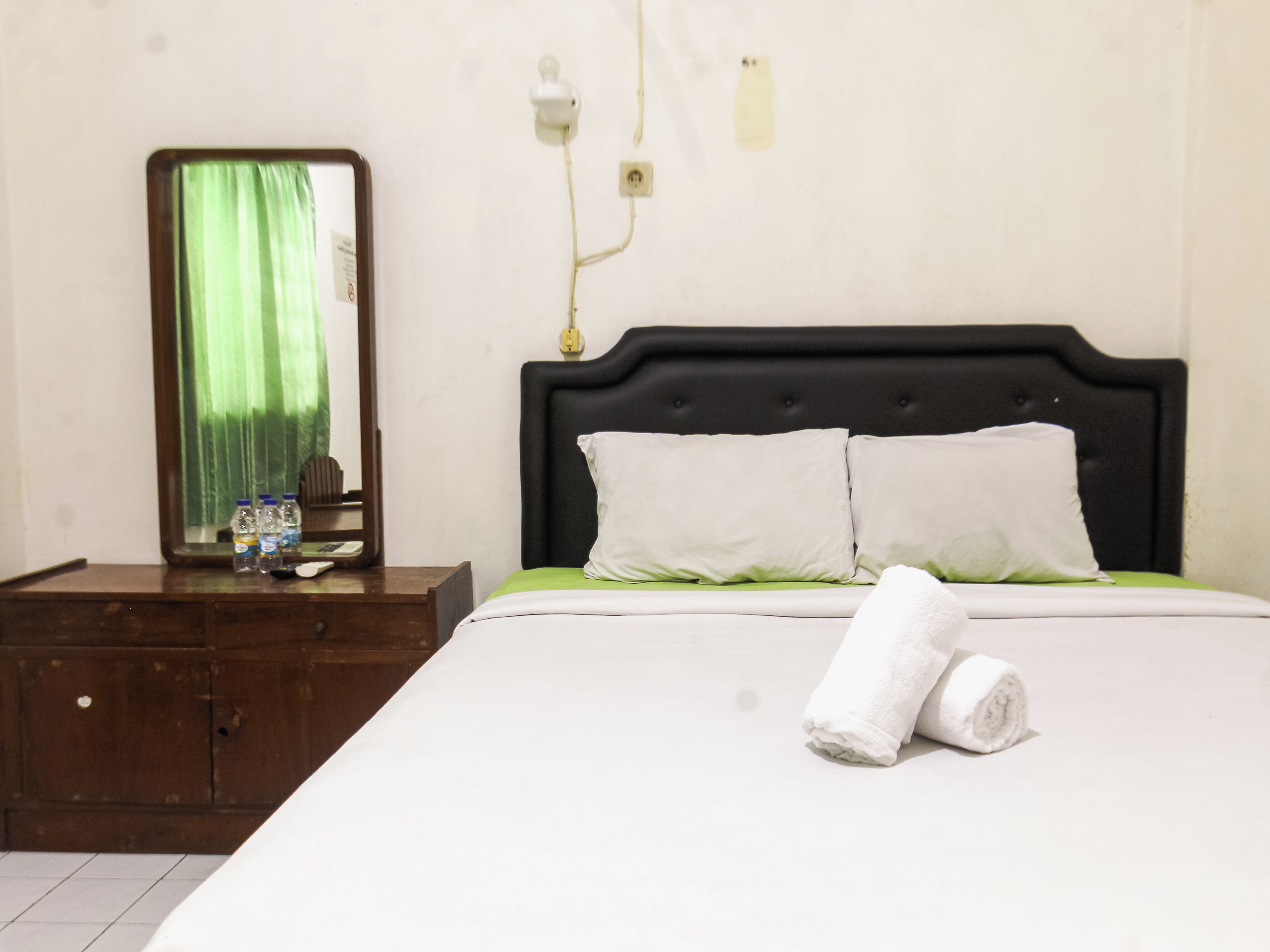Un letto rifatto con biancheria bianca e due cuscini, un comodino in legno con uno specchio e alcuni oggetti, e un lampadario fissato al muro.
