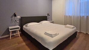 A neatly made bed with white linens and a folded towel, a bedside table with a lamp, and a window with sheer curtains. - Cozy apartment near the center, excellent for families (Bardolino)