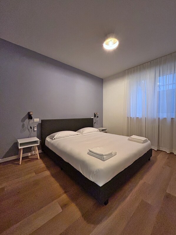 Uma cama bem arrumada com lençóis brancos e uma toalha dobrada, uma mesinha de cabeceira com abajur, e uma janela com cortinas finas.