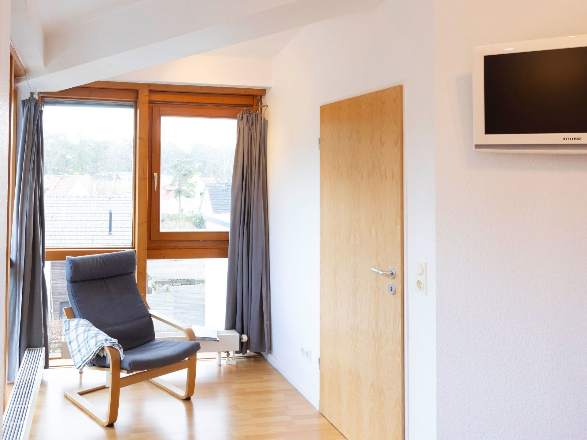 Una habitación moderna con piso de madera, una silla gris, una puerta de madera y un televisor fijado a la pared.