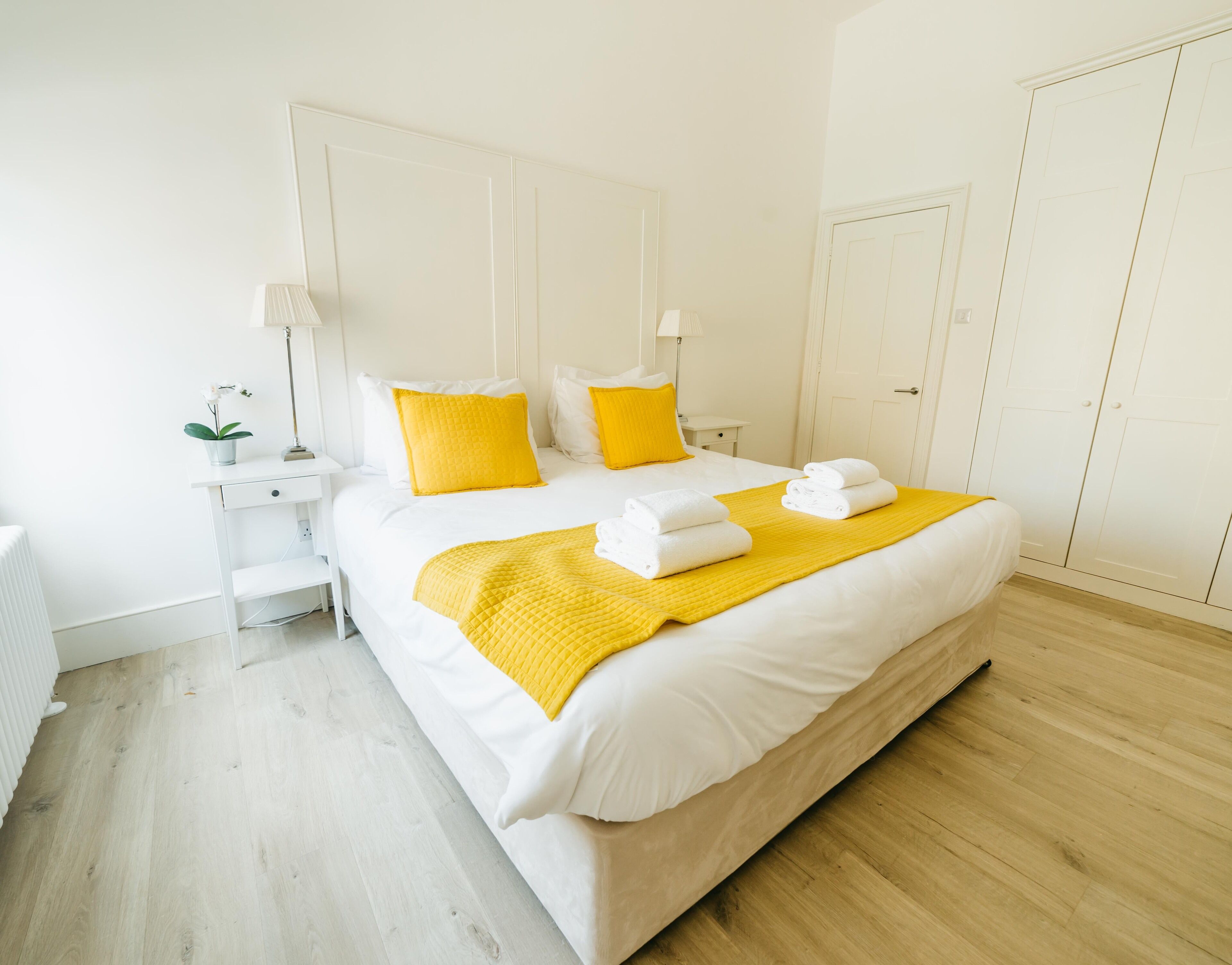 A neatly arranged hotel room with a large bed, white bedding, and yellow accents.