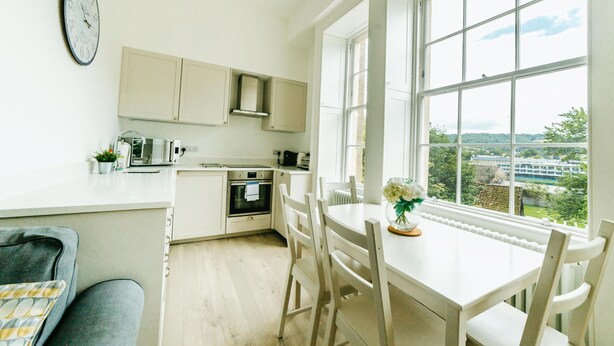 A kitchen with a view of a green landscape, a dining table with chairs, and a clock on the wall. - Argyle St Apartments  (Bath)
