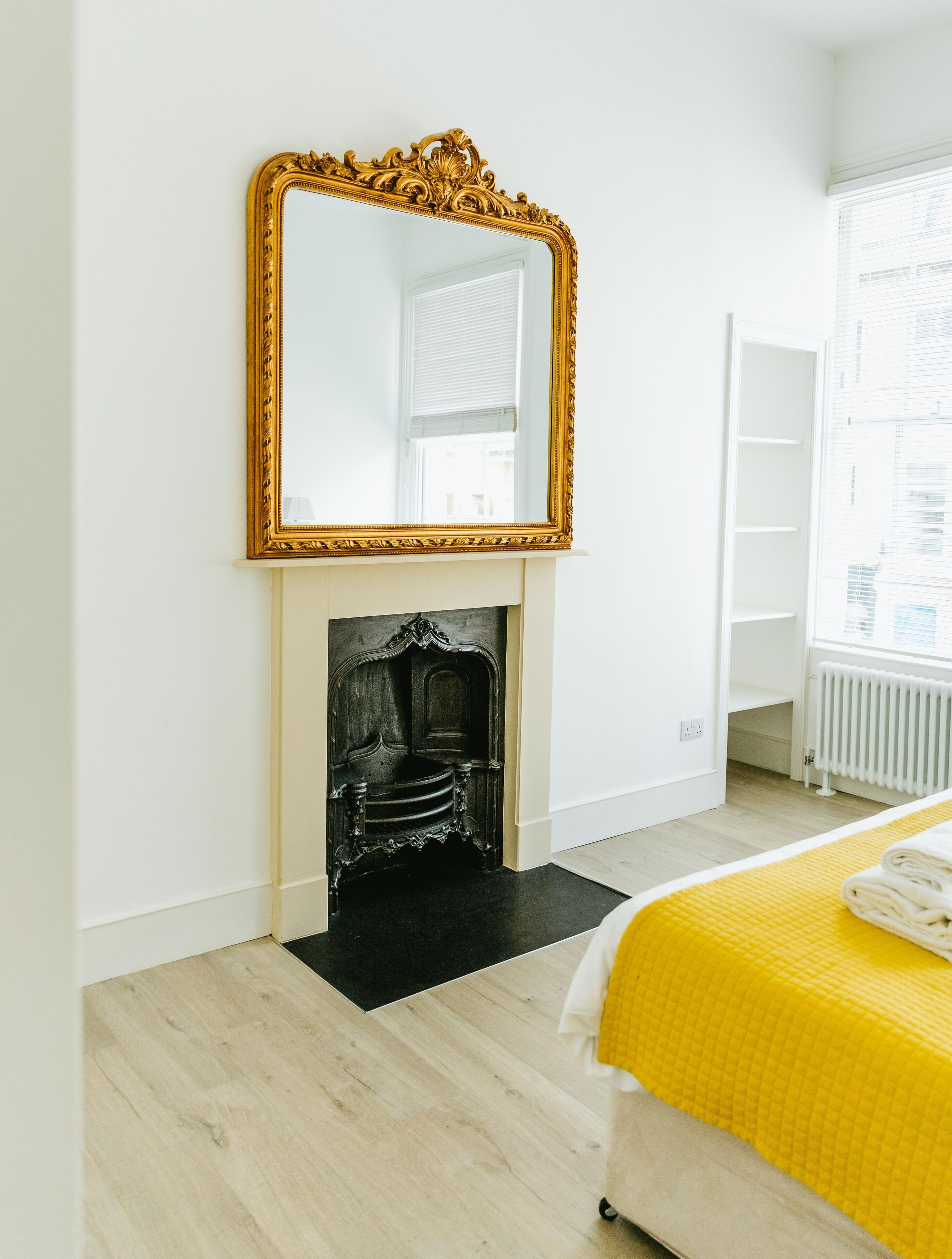 A bedroom with a large ornate mirror, a fireplace, a bed with a yellow bedspread, and a white radiator.