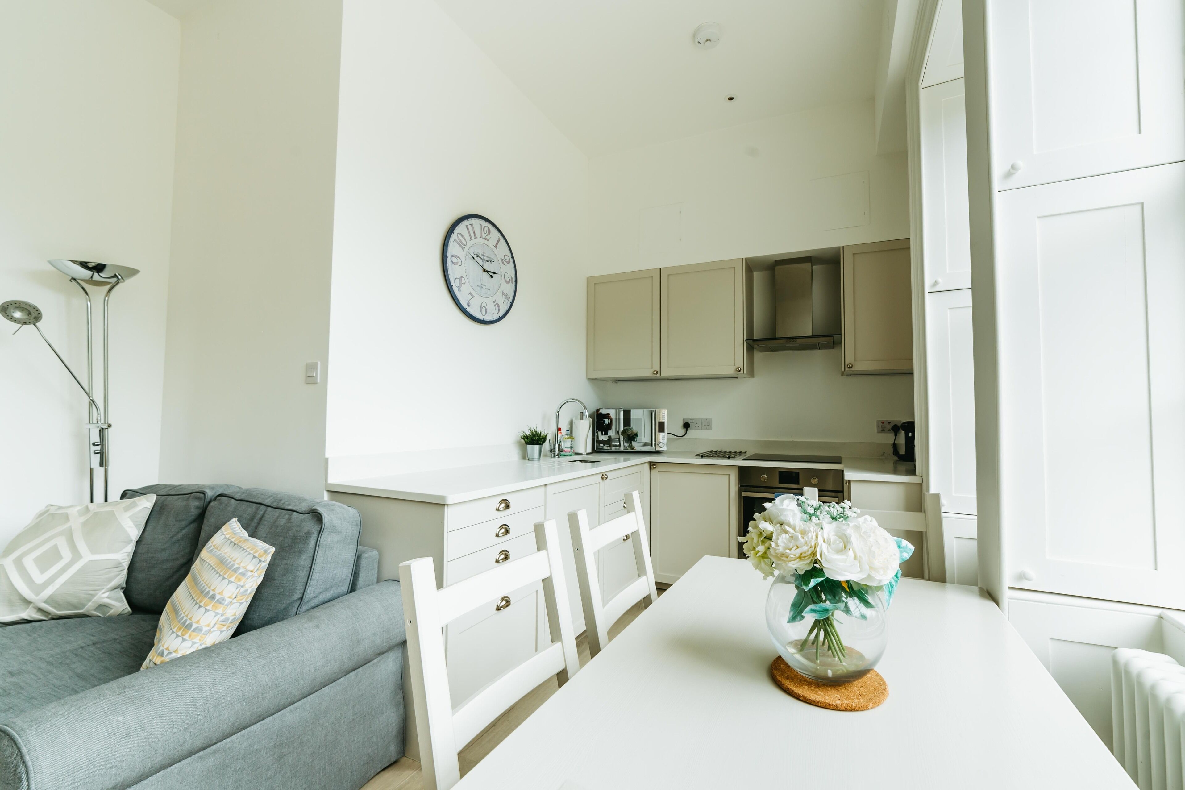 Een moderne keuken met witte kastjes, een eettafel met stoelen en een klok aan de muur.