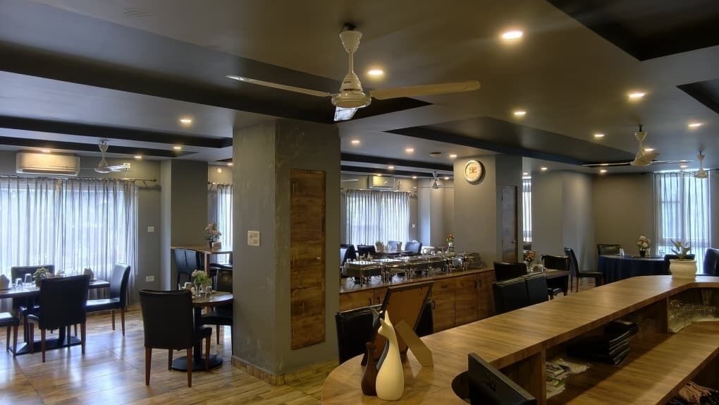 A modern hotel lobby with a long wooden reception desk, dining area with chairs and tables, a bar area with a counter, and ceiling fans.