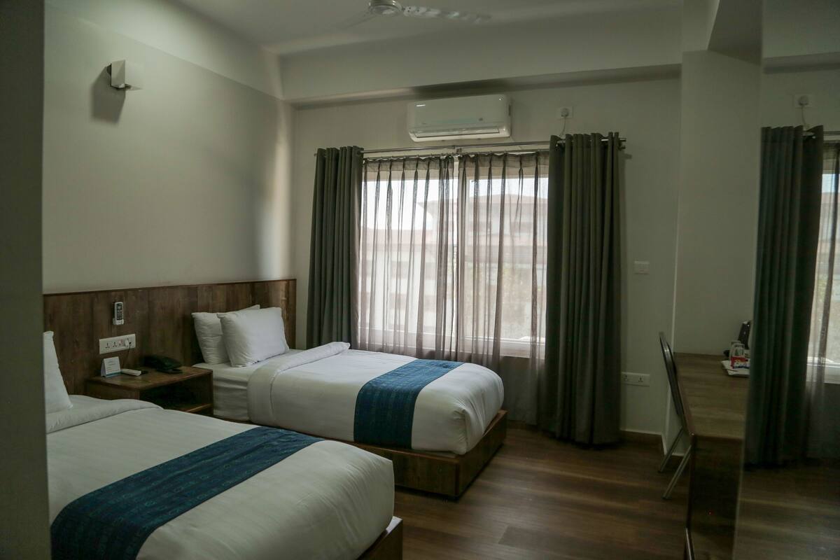 Habitación de hotel con dos camas, cabecero de madera, mesita de noche, equipo de aire acondicionado y vistas al exterior a través de cortinas translúcidas.