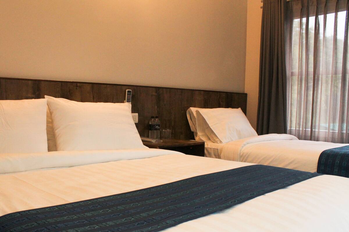 Habitación de hotel con dos camas, cabecera de madera, mesita de noche con una botella y ventana con cortinas.