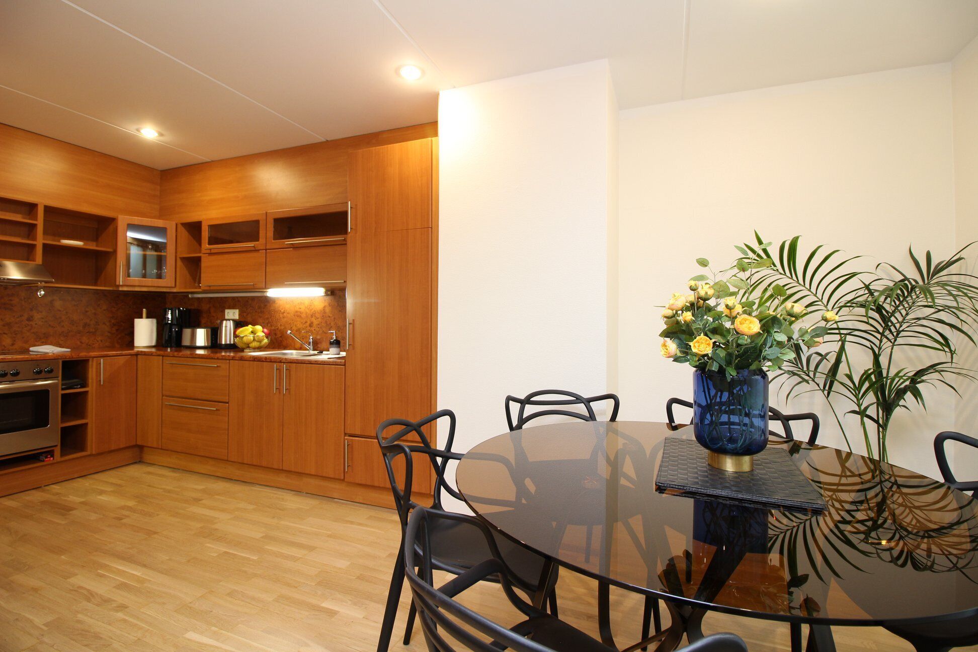 A modern kitchen with wooden cabinets, a stainless steel oven, and a dining area with a glass table and chairs.