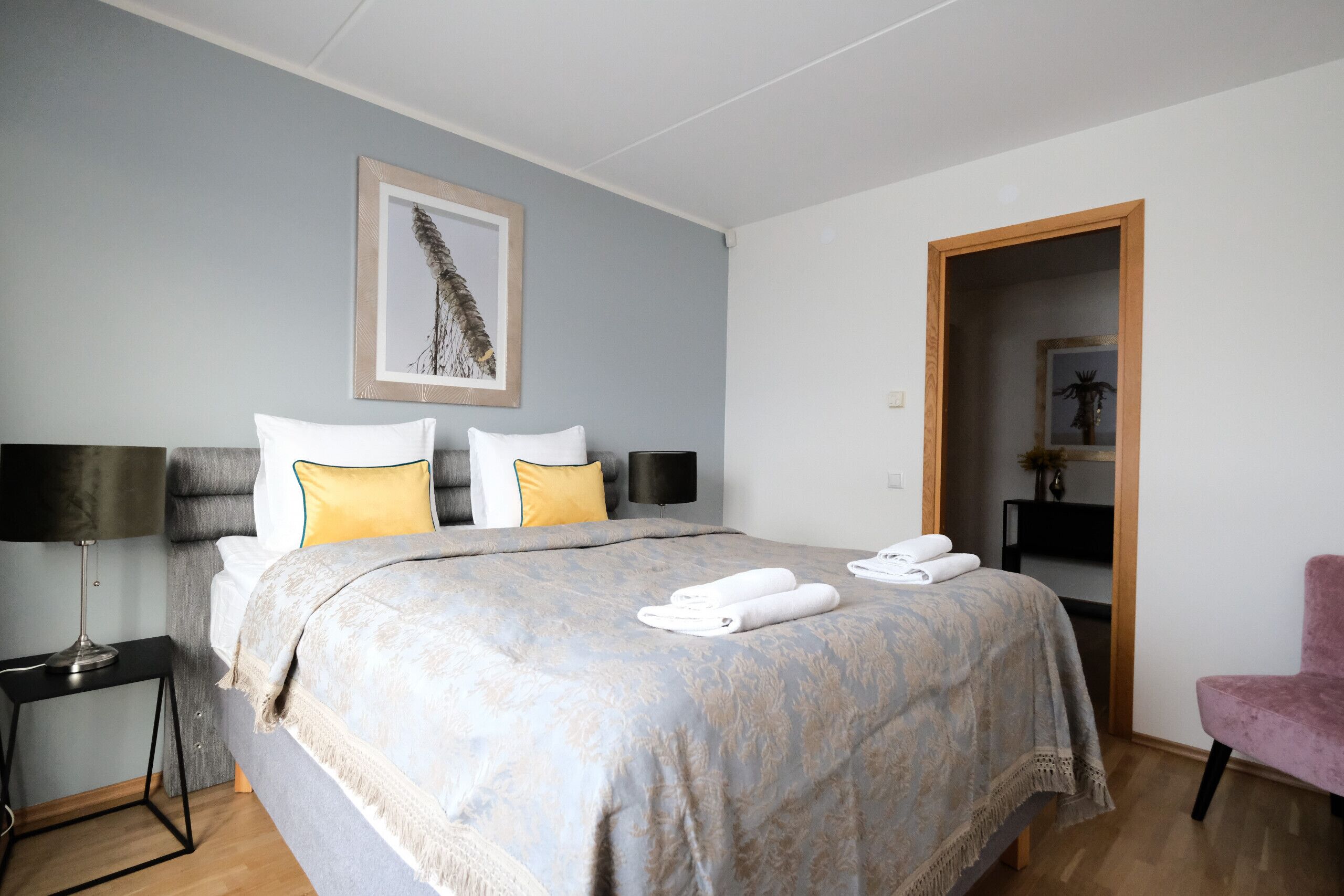 A neatly made bed with a patterned bedspread, two pillows, and a pair of folded towels. A framed artwork hangs above the bed, and a chair is visible to the right.